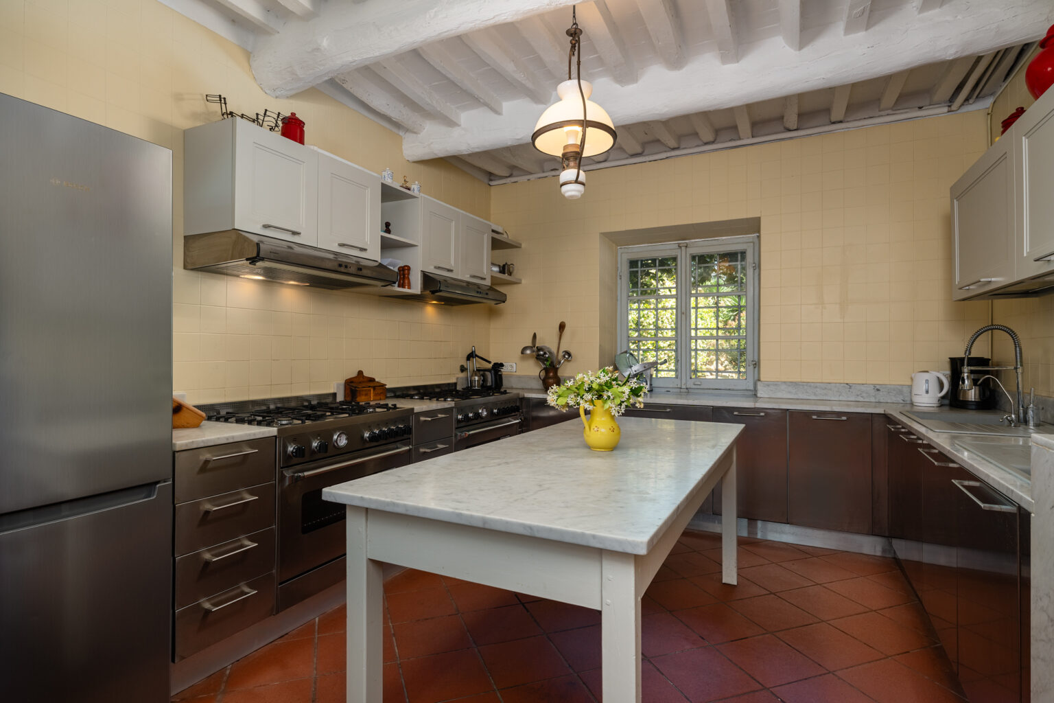 Modern kitchen interior featuring stainless steel appliances, a marble countertop island, and a charming yellow vase with flowers. The space has wooden beams, tiled flooring, and ample natural light from a window, creating a warm and inviting atmosphere. | Only on SopranoVillas