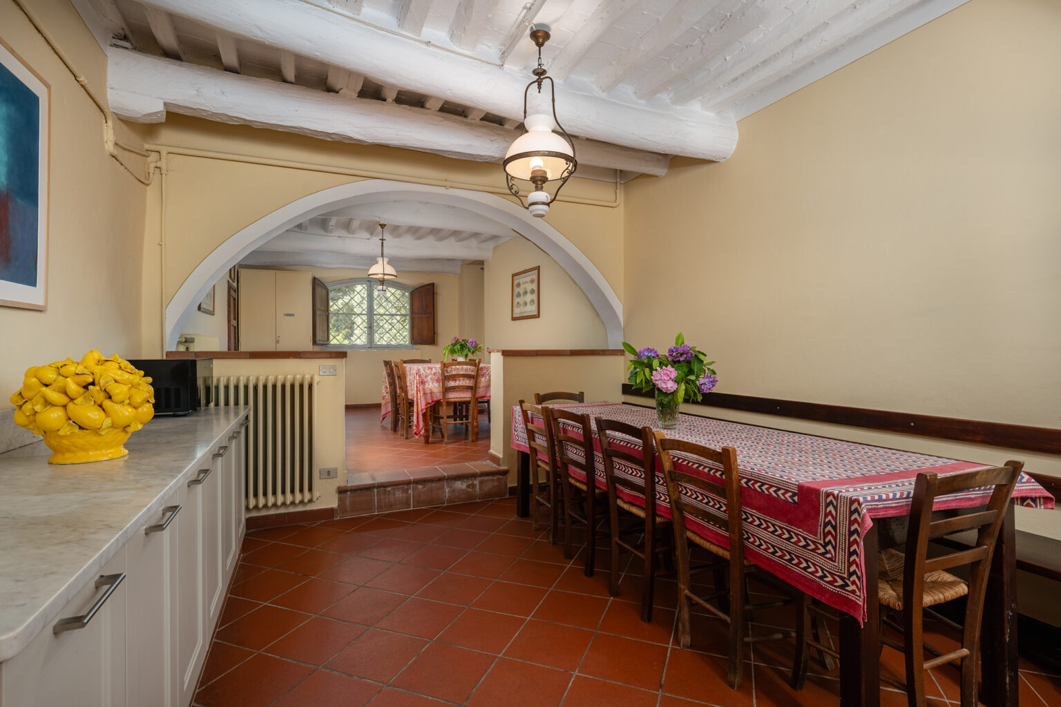 Spacious kitchen dining area featuring a long wooden table with patterned tablecloth, surrounded by chairs, and an arched entryway. Decorated with a vibrant yellow ceramic fruit bowl and fresh flowers, the room has whitewashed beams and terracotta tile flooring, creating a warm and inviting atmosphere. | Only on SopranoVillas