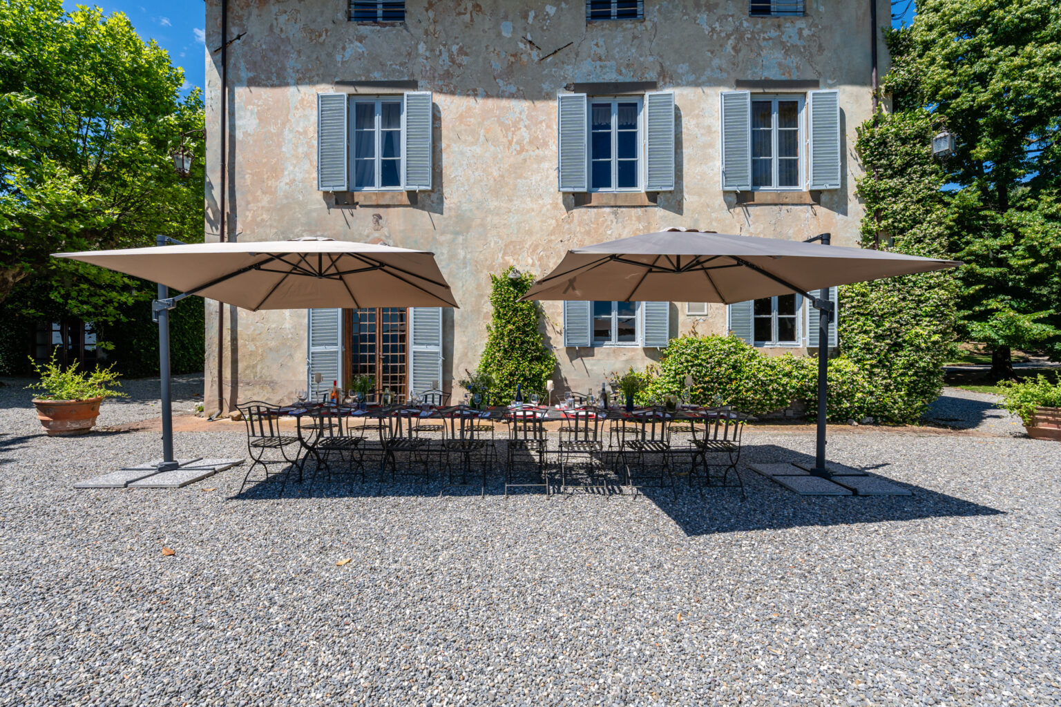 Outdoor dining area with shaded umbrellas in front of a rustic stone building, surrounded by greenery and gravel pathway. Perfect for gatherings and events. | Only on SopranoVillas