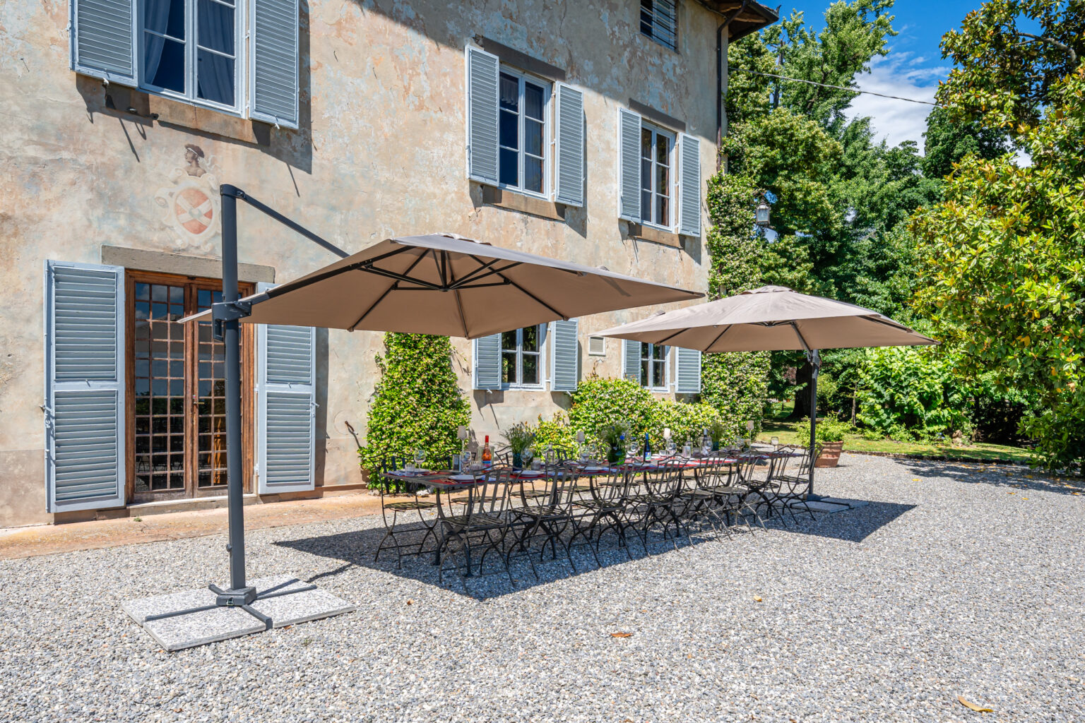 Outdoor dining area featuring two large umbrellas over a long table set for a meal, with a rustic house in the background and lush greenery surrounding the space. Ideal for gatherings and al fresco dining. | Only on SopranoVillas