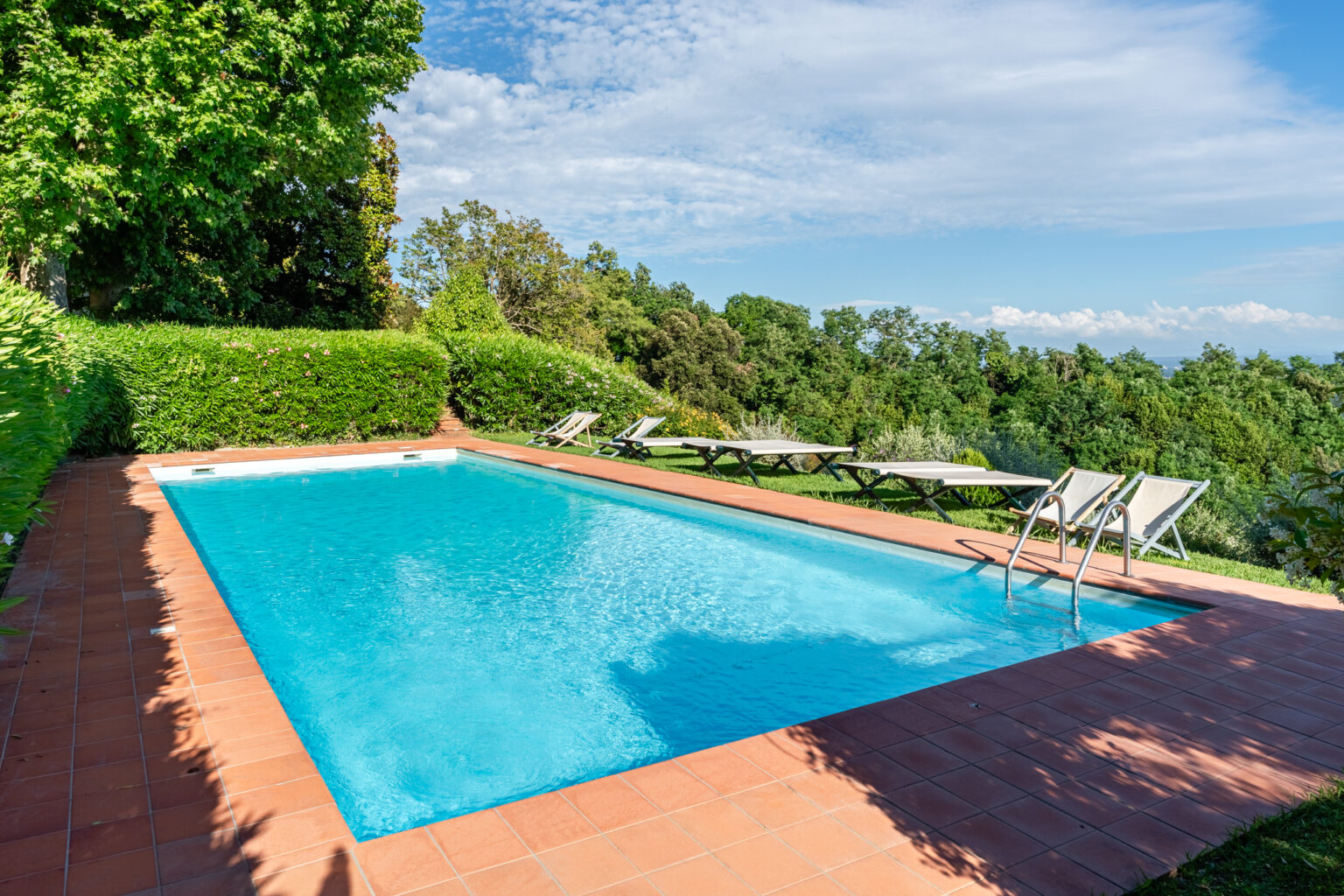 A serene outdoor swimming pool surrounded by lush greenery and sun loungers, set against a backdrop of blue skies and distant mountains. Ideal for relaxation and leisure activities. | Only on SopranoVillas