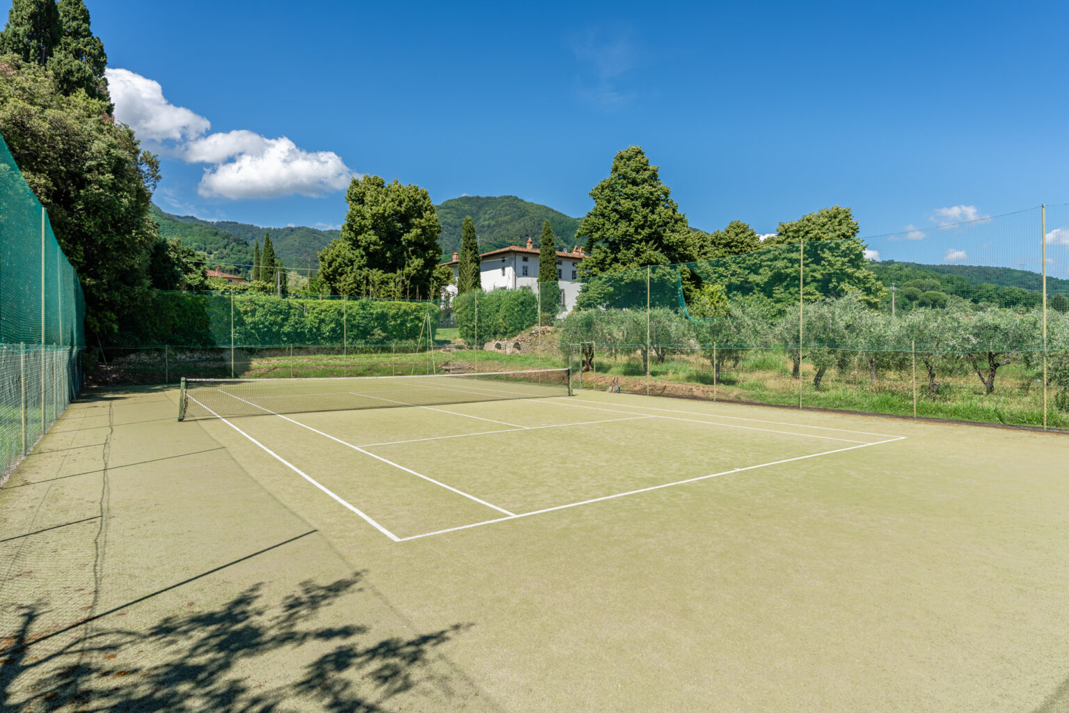 Tennis court surrounded by lush greenery and mountains under a clear blue sky, featuring a sandy surface and white boundary lines. | Only on SopranoVillas