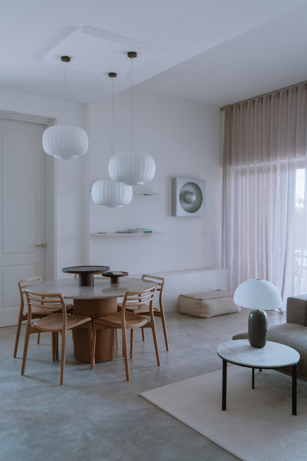 Modern dining area featuring a round wooden table, four wooden chairs, and stylish pendant lights. The space is adorned with light curtains, a minimalist wall art piece, and a soft glowing lamp, creating a serene and inviting atmosphere. | Only on SopranoVillas