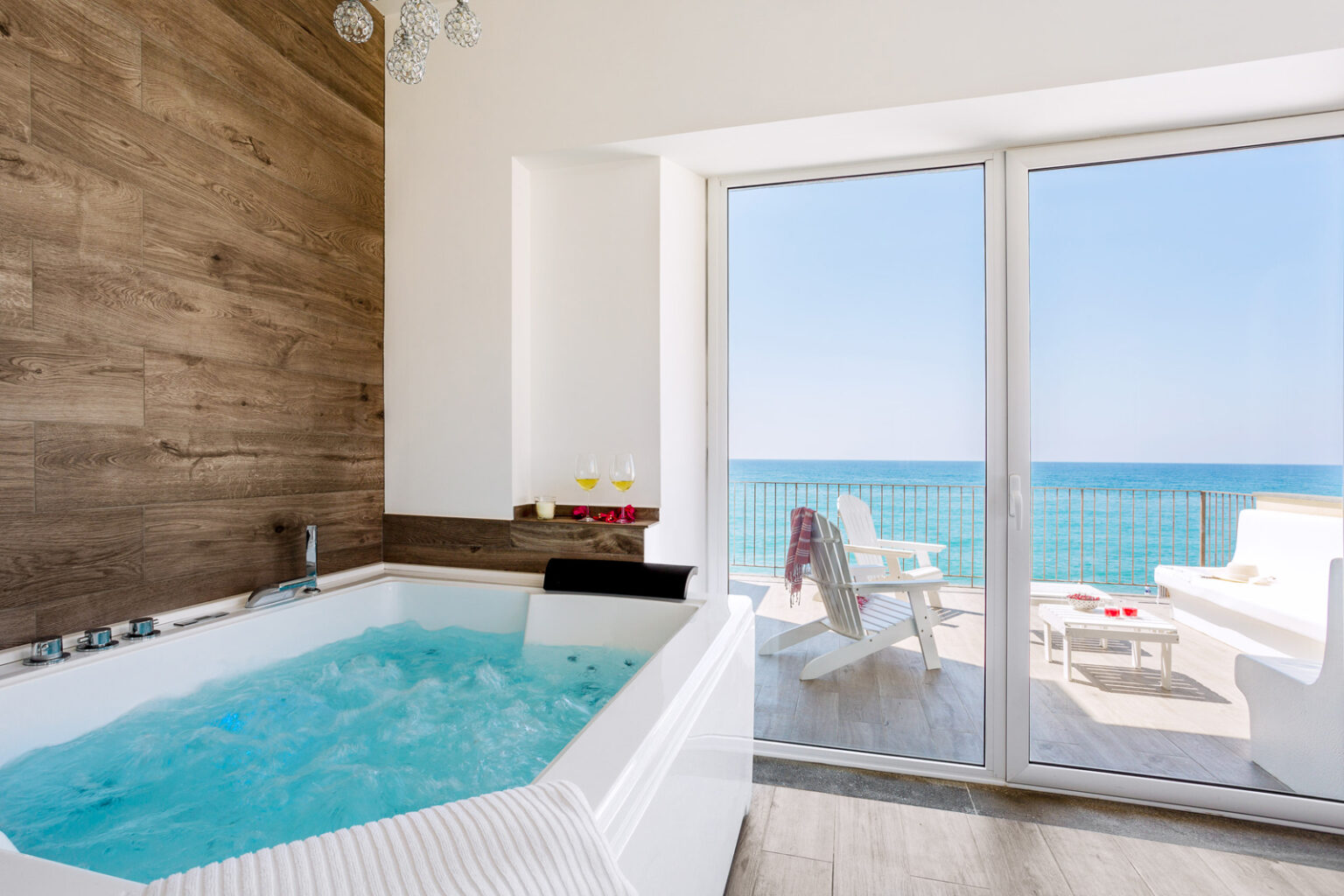 Luxurious bathroom with a modern hot tub overlooking the ocean, featuring large glass doors that open to a scenic balcony with white lounge chairs and a beautiful sea view. | Only on SopranoVillas