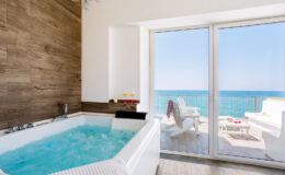 Luxurious bathroom with a modern hot tub overlooking the ocean, featuring large glass doors that open to a scenic balcony with white lounge chairs and a beautiful sea view.