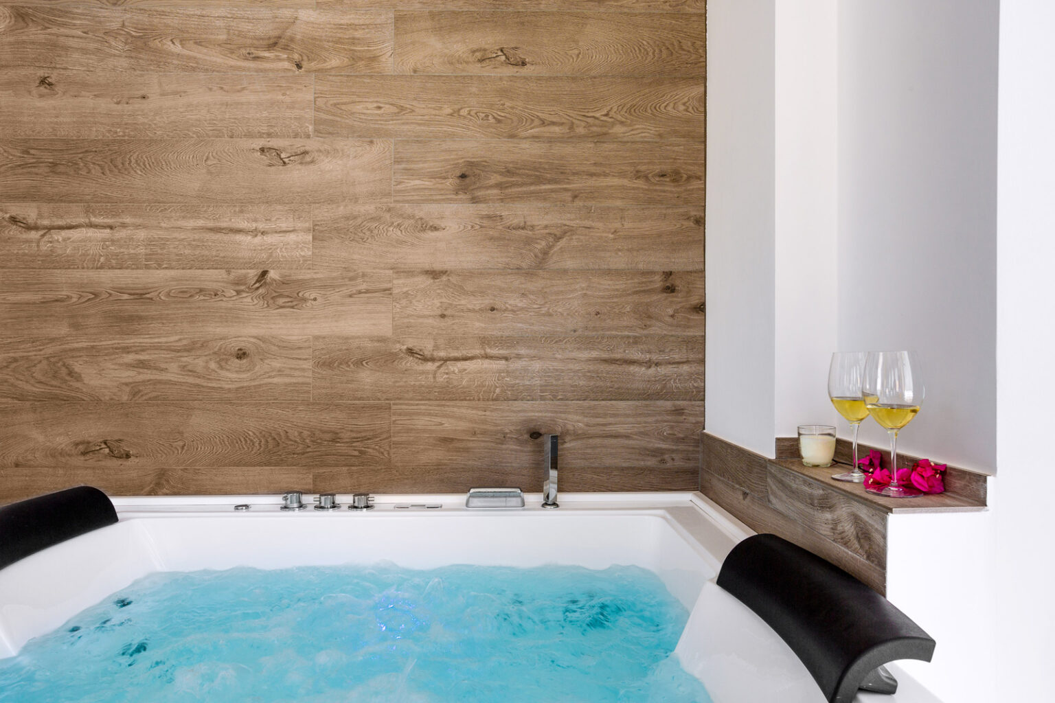 Modern bathroom featuring a luxurious jacuzzi tub with blue water, surrounded by a stylish wooden accent wall. Nearby, two glasses of champagne and pink flowers add a touch of elegance. Perfect for relaxation and spa-like experiences. | Only on SopranoVillas