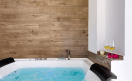 Modern bathroom featuring a luxurious jacuzzi tub with blue water, surrounded by a stylish wooden accent wall. Nearby, two glasses of champagne and pink flowers add a touch of elegance. Perfect for relaxation and spa-like experiences.