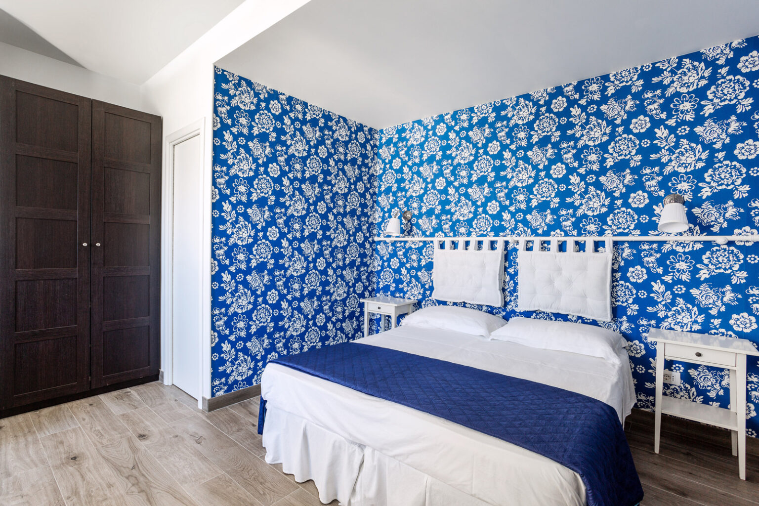 Cozy bedroom featuring vibrant blue floral wallpaper, a white bed with a navy blue throw, and elegant white bedside tables, complemented by a dark wooden wardrobe. Ideal for a refreshing and stylish retreat. | Only on SopranoVillas