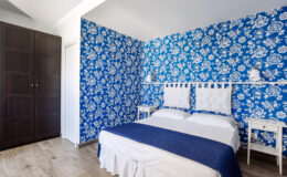 Cozy bedroom featuring vibrant blue floral wallpaper, a white bed with a navy blue throw, and elegant white bedside tables, complemented by a dark wooden wardrobe. Ideal for a refreshing and stylish retreat.