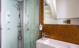 Modern bathroom featuring a glass shower enclosure, sleek white bathtub, and vibrant orange mosaic tile wall. The space is well-lit with a minimalist design and contemporary fixtures.