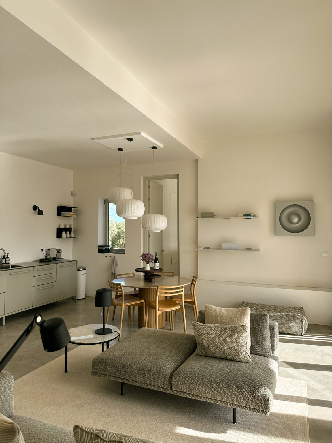 Modern kitchen and dining area featuring a wooden table with chairs, pendant lighting, and minimalist decor. The space includes a sleek kitchen with cabinetry, a window for natural light, and decorative shelving on the wall. | Only on SopranoVillas