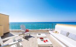 Cozy seaside balcony featuring white outdoor furniture, a stunning ocean view, and refreshing drinks, perfect for relaxation and enjoying the beach atmosphere.