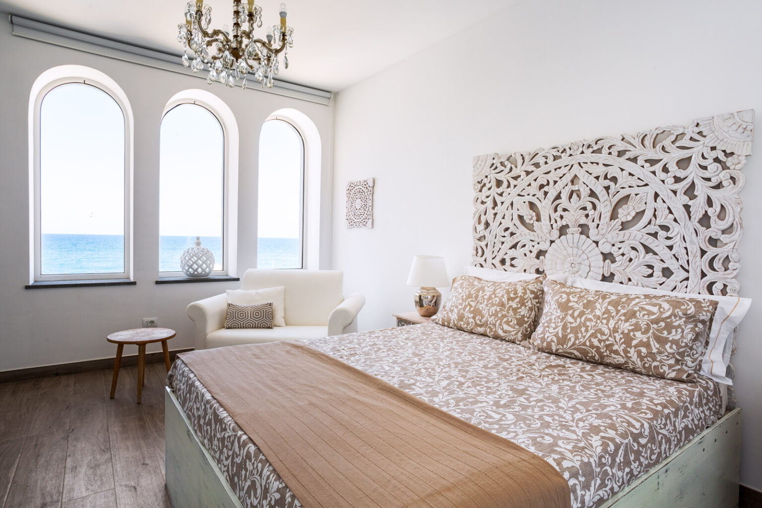Bright and airy bedroom featuring a large bed with decorative brown and beige bedding, a stylish carved headboard, and a cozy seating area. The room has three arched windows offering a stunning ocean view, complemented by elegant chandelier lighting and modern decor. | Only on SopranoVillas