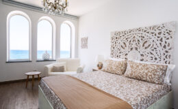 Bright and airy bedroom featuring a large bed with decorative brown and beige bedding, a stylish carved headboard, and a cozy seating area. The room has three arched windows offering a stunning ocean view, complemented by elegant chandelier lighting and modern decor.
