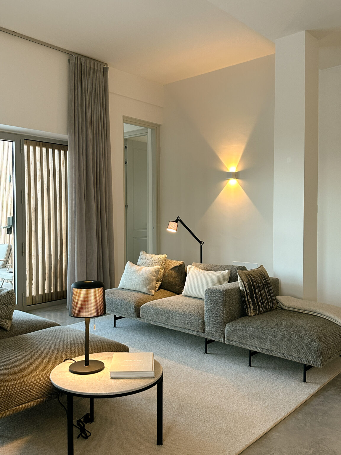 Modern living room featuring a gray sectional sofa with decorative pillows, a stylish floor lamp, and soft lighting. Large windows with sheer curtains allow natural light, enhancing the cozy atmosphere. | Only on SopranoVillas