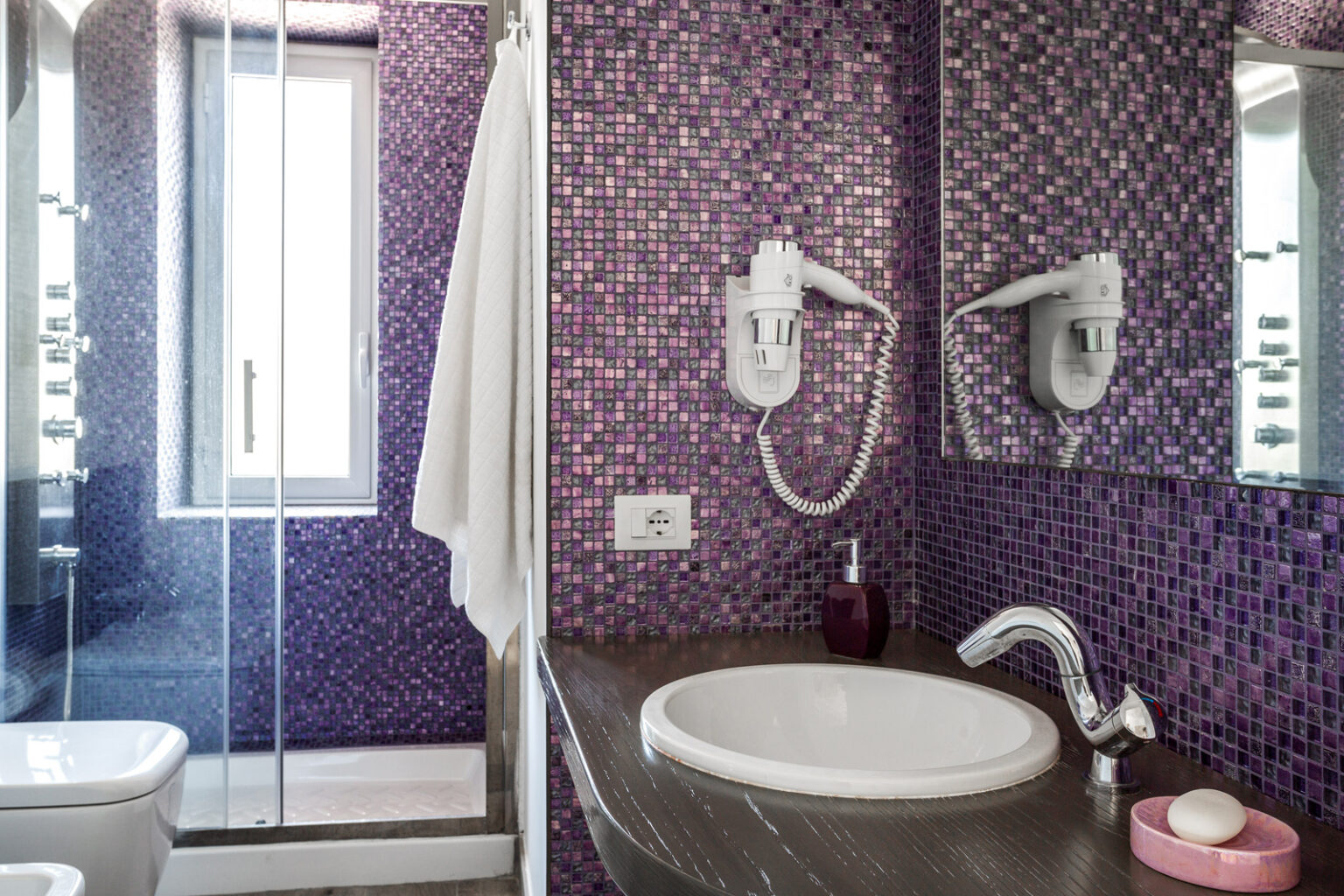Modern bathroom featuring purple mosaic tiles, a white sink with a chrome faucet, a shower area with glass doors, and a towel hanging on the wall. The design combines functionality with vibrant aesthetics. | Only on SopranoVillas