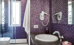 Modern bathroom featuring purple mosaic tiles, a white sink with a chrome faucet, a shower area with glass doors, and a towel hanging on the wall. The design combines functionality with vibrant aesthetics.