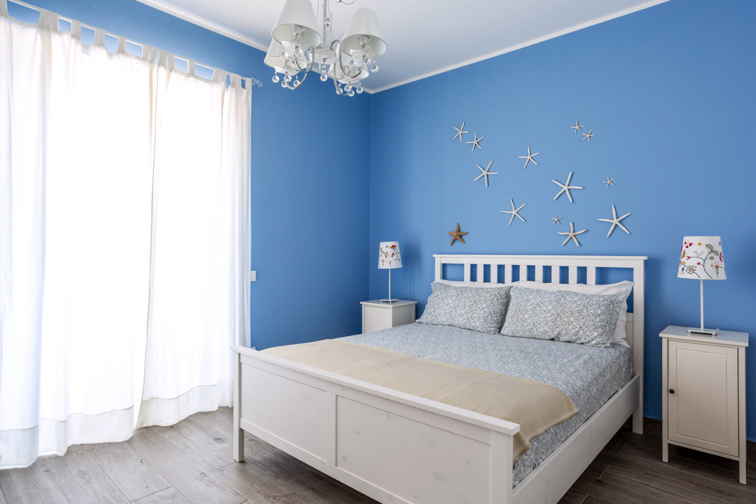 Bright and airy bedroom featuring a blue accent wall, a white wooden bed with light blue bedding, and decorative starfish on the wall. The room includes a nightstand with a lamp and large windows with sheer curtains, creating a serene atmosphere. | Only on SopranoVillas