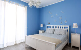 Bright and airy bedroom featuring a blue accent wall, a white wooden bed with light blue bedding, and decorative starfish on the wall. The room includes a nightstand with a lamp and large windows with sheer curtains, creating a serene atmosphere.