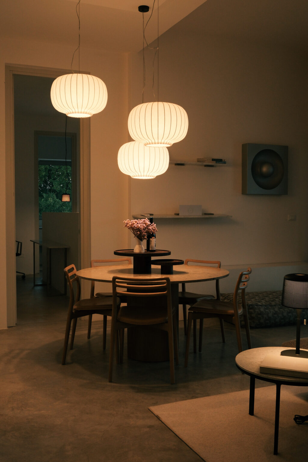 Modern dining area featuring a round wooden table, stylish chairs, and a vase of flowers, illuminated by contemporary pendant lighting, with a minimalist decor in soft tones. | Only on SopranoVillas