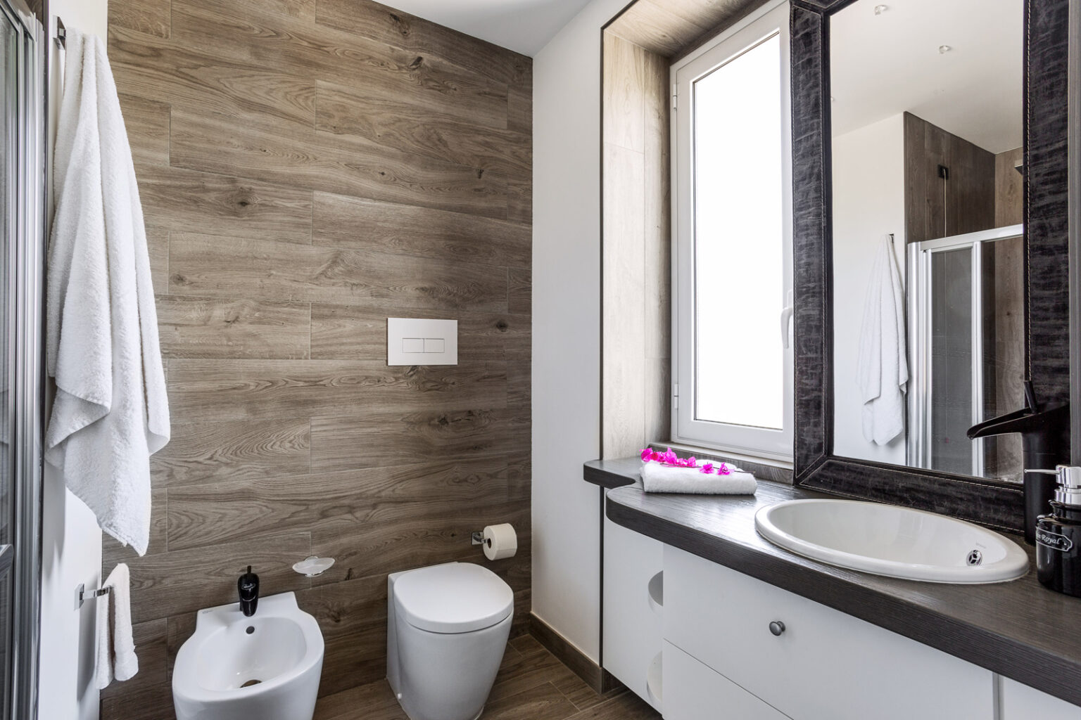 Modern bathroom interior featuring wooden wall panels, a white toilet, bidet, and a stylish sink with a round mirror. Natural light enters through a window, enhancing the contemporary design with a touch of elegance. Soft towels and decorative flowers add a cozy ambiance. | Only on SopranoVillas