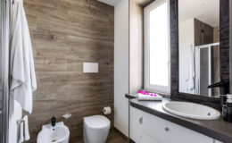 Modern bathroom interior featuring wooden wall panels, a white toilet, bidet, and a stylish sink with a round mirror. Natural light enters through a window, enhancing the contemporary design with a touch of elegance. Soft towels and decorative flowers add a cozy ambiance.