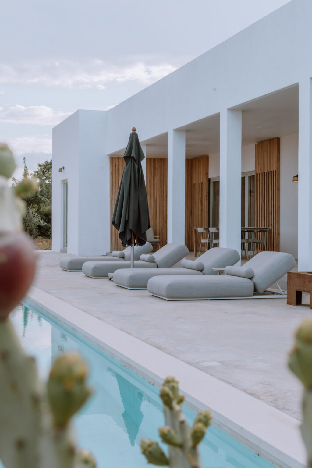 Modern outdoor lounge area featuring gray chaise lounges and a large umbrella beside a swimming pool, with a backdrop of minimalist architecture and wooden accents. | Only on SopranoVillas