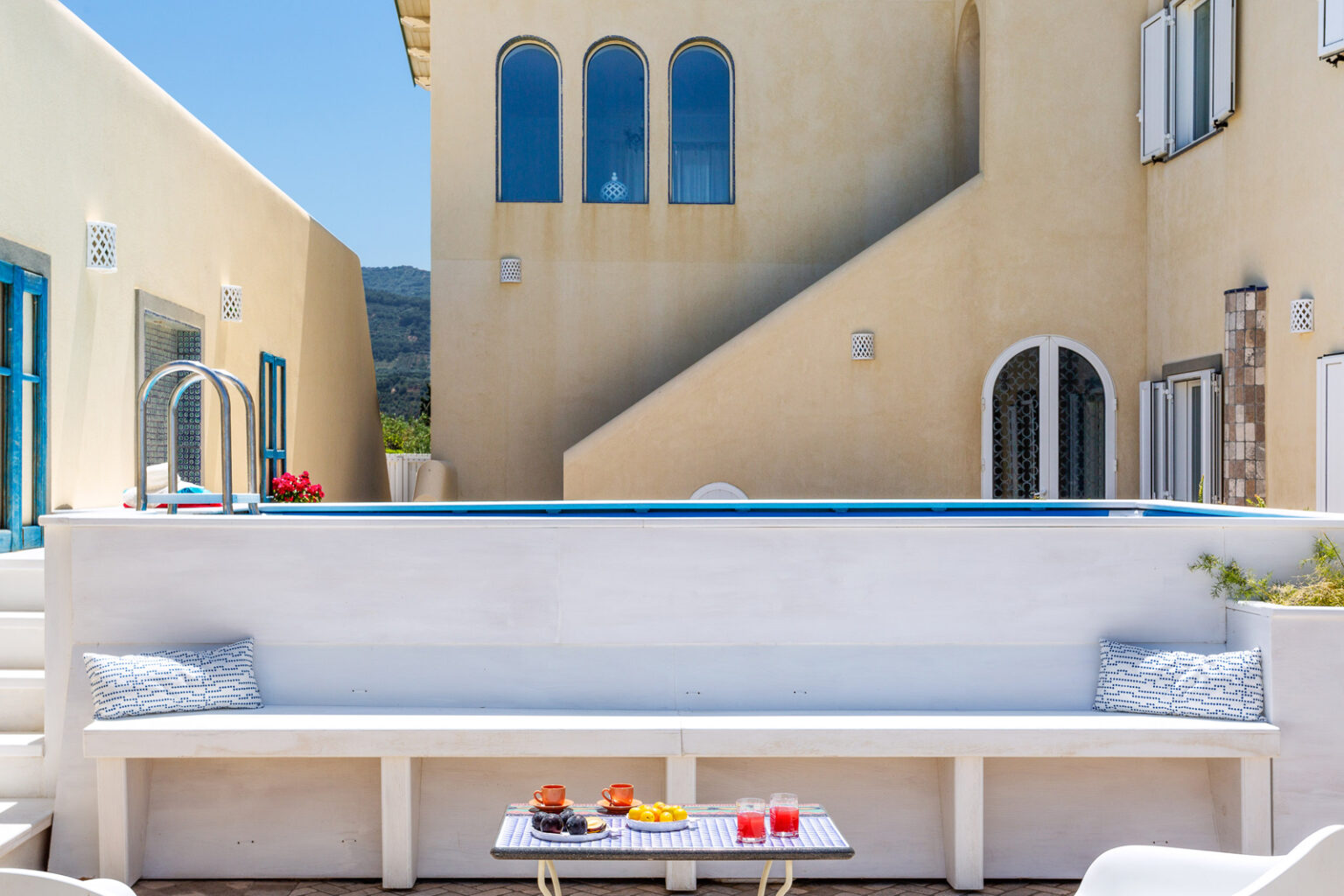 Modern outdoor seating area with a white bench, colorful drinks on a table, and a backdrop of a Mediterranean-style building with blue windows and a pool, surrounded by a sunny landscape. | Only on SopranoVillas