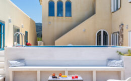 Modern outdoor seating area with a white bench, colorful drinks on a table, and a backdrop of a Mediterranean-style building with blue windows and a pool, surrounded by a sunny landscape.