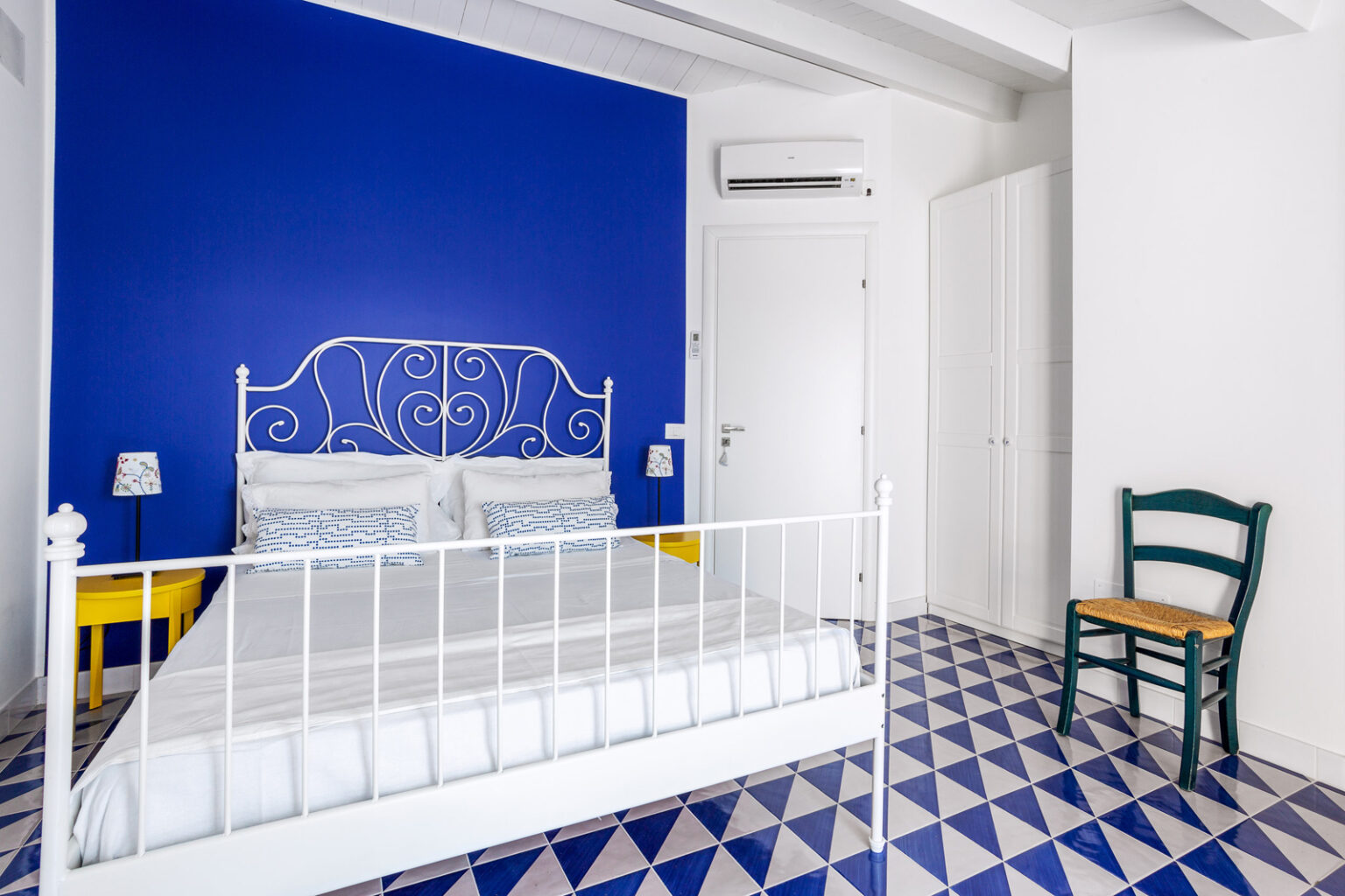 Bright and airy bedroom featuring a white metal bed, vibrant blue accent wall, and geometric patterned floor tiles. The room includes a small green chair and bedside lamps, creating a cozy and inviting atmosphere. Ideal for a relaxing getaway or stylish home decor inspiration. | Only on SopranoVillas
