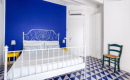 Bright and airy bedroom featuring a white metal bed, vibrant blue accent wall, and geometric patterned floor tiles. The room includes a small green chair and bedside lamps, creating a cozy and inviting atmosphere. Ideal for a relaxing getaway or stylish home decor inspiration.
