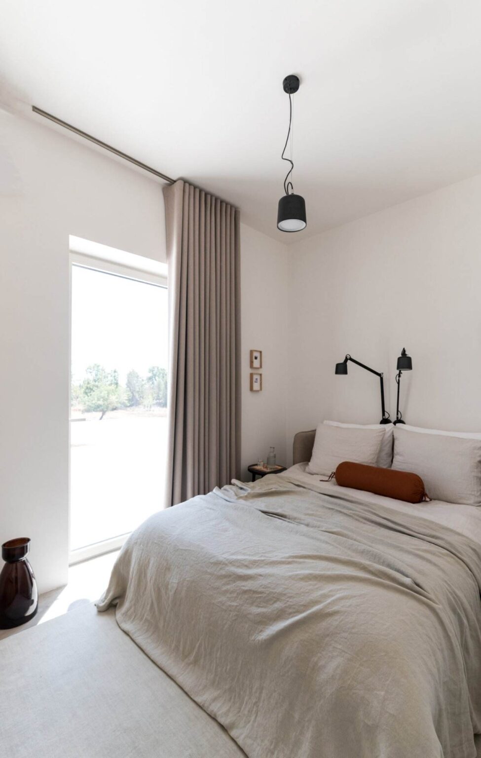 Modern bedroom interior featuring a neatly made bed with white linens and an orange accent pillow, complemented by minimalist decor, wall-mounted reading lights, and floor-to-ceiling curtains, with a view of nature through a large window. | Only on SopranoVillas