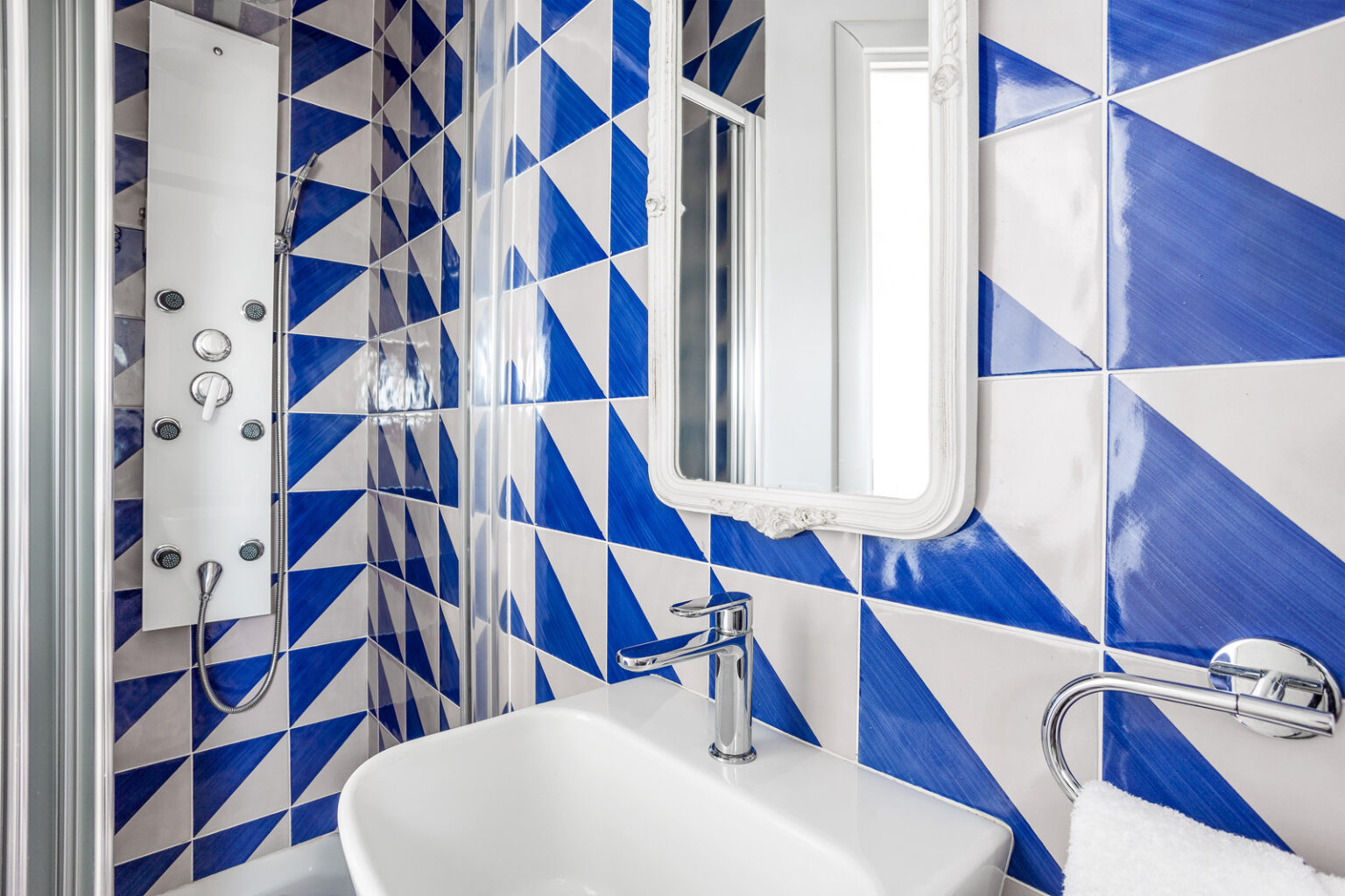 Modern bathroom featuring a stylish shower with a sleek panel and multiple showerhead options, complemented by a vibrant blue and white geometric tile design. A contemporary sink with a chrome faucet and a decorative mirror enhance the space's aesthetic appeal. | Only on SopranoVillas