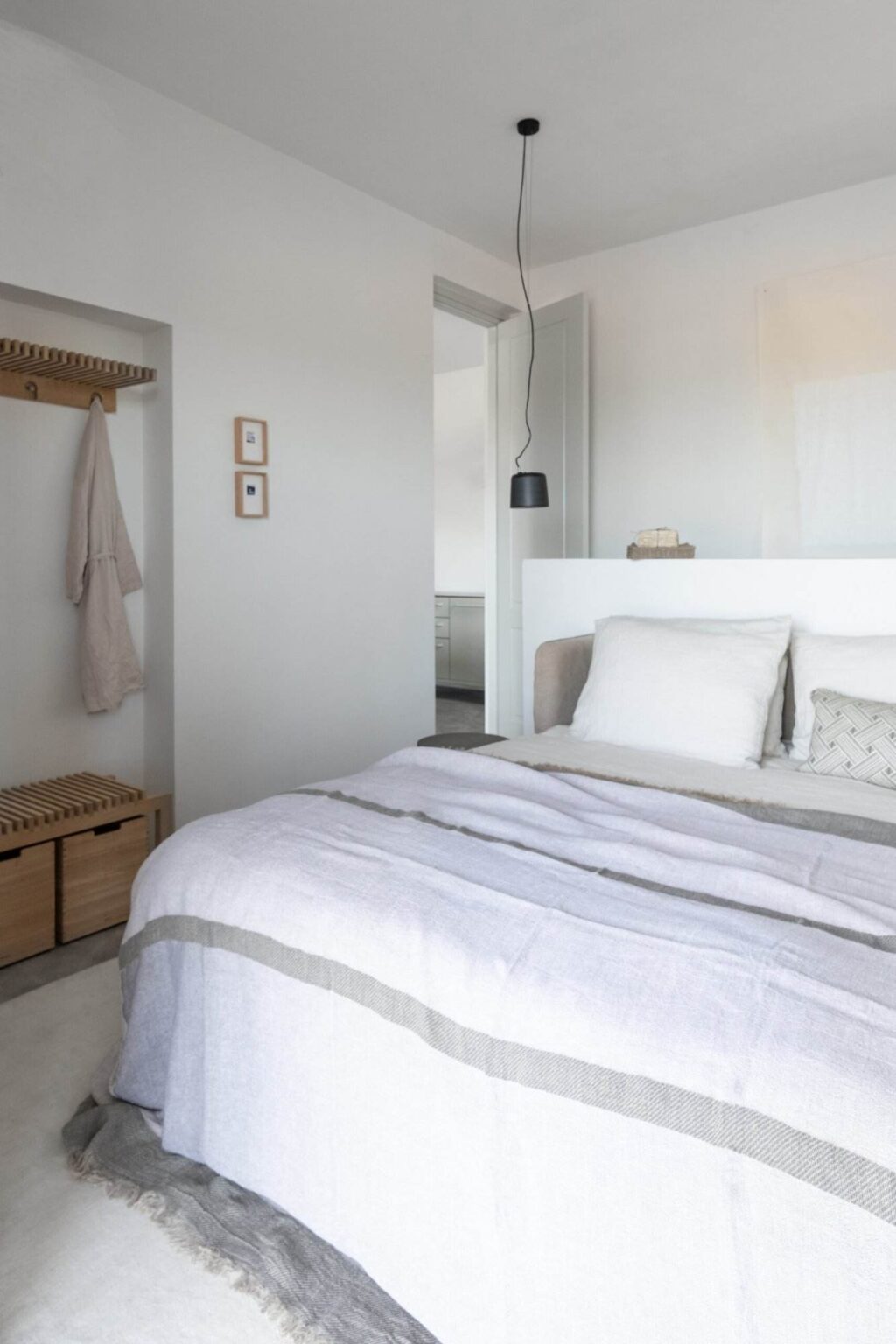 Modern bedroom interior featuring a cozy bed with a light gray blanket, decorative pillows, and a minimalist design. The room includes a wooden bench with storage, a hanging coat rack, and a sleek pendant light, all set against neutral-toned walls for a calming atmosphere. | Only on SopranoVillas