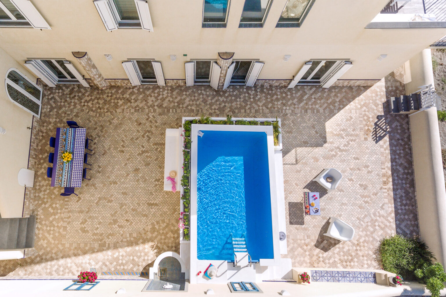 Aerial view of a modern outdoor patio featuring a rectangular swimming pool, surrounded by potted plants and stylish seating. A colorful dining table is set for outdoor meals, highlighting the inviting atmosphere of this luxurious outdoor space. | Only on SopranoVillas