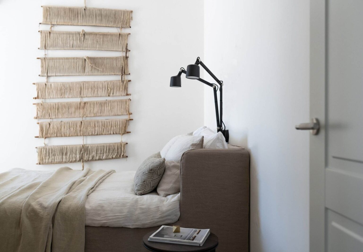 Cozy bedroom featuring a minimalist design, with a neutral color palette. A comfortable bed is adorned with soft linens and decorative pillows, while a stylish black reading lamp is mounted beside the bed. A unique wall hanging made of woven fibers adds texture to the white wall, enhancing the serene atmosphere of the space. A small side table holds a magazine, completing the inviting look. | Only on SopranoVillas