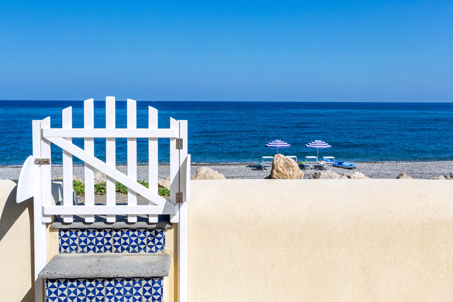 White wooden gate leading to a serene beach with clear blue waters, sun loungers, and umbrellas under a bright sky. Ideal for vacation and relaxation. | Only on SopranoVillas