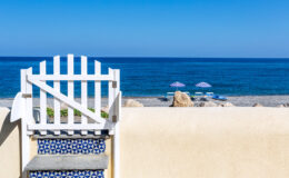 White wooden gate leading to a serene beach with clear blue waters, sun loungers, and umbrellas under a bright sky. Ideal for vacation and relaxation.