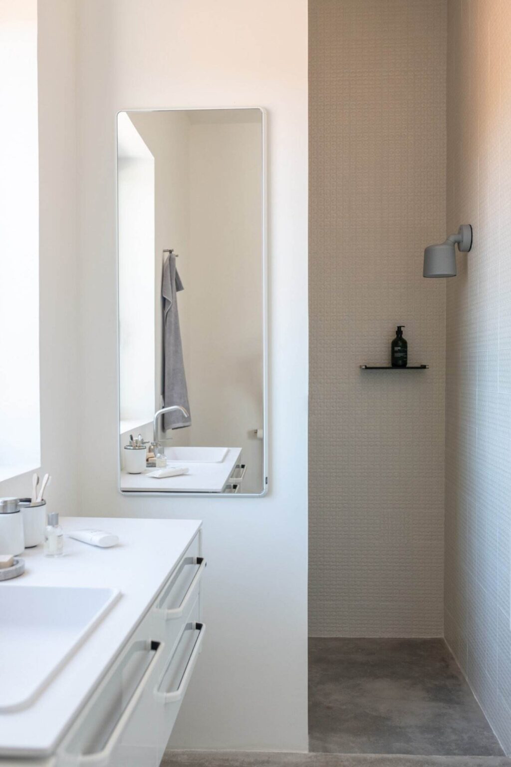 Modern bathroom interior featuring a sleek white vanity with a mirror, neatly arranged toiletries, and a shower area with textured wall tiles. A gray towel hangs on a rod, and a minimalist light fixture illuminates the space, creating a clean and inviting atmosphere. | Only on SopranoVillas