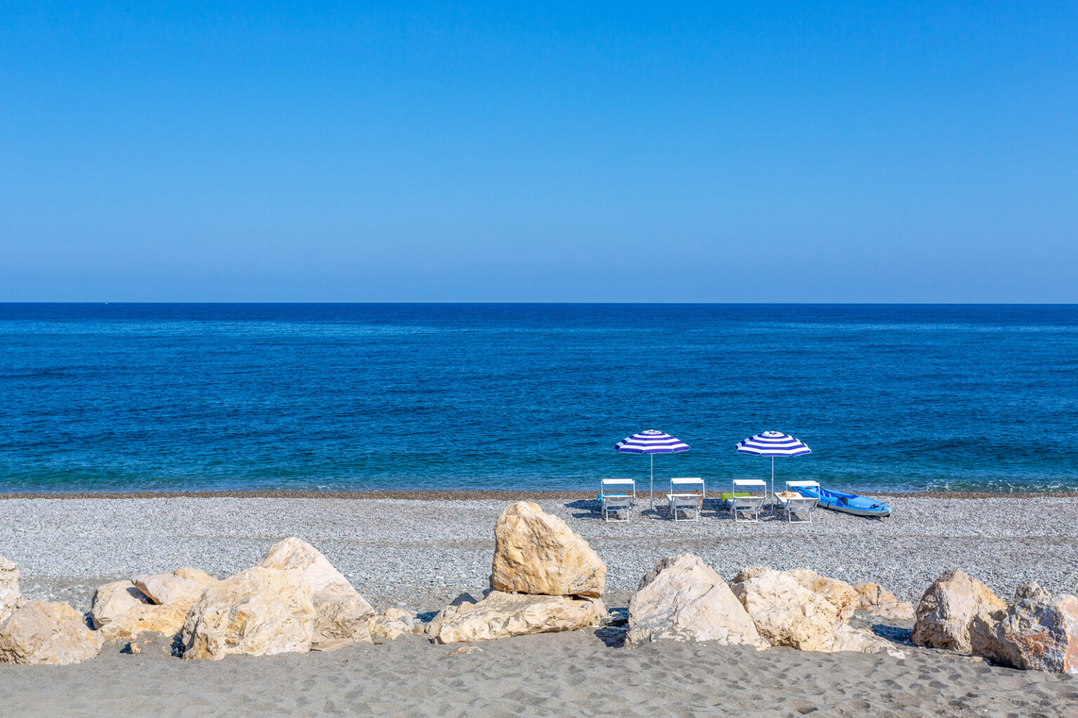 Tranquil beach scene featuring two purple-striped umbrellas and lounge chairs on a sandy shore, with clear blue ocean waves and a bright sky in the background. Perfect for vacation and relaxation imagery. | Only on SopranoVillas