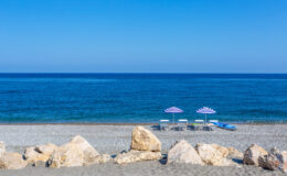 Tranquil beach scene featuring two purple-striped umbrellas and lounge chairs on a sandy shore, with clear blue ocean waves and a bright sky in the background. Perfect for vacation and relaxation imagery.