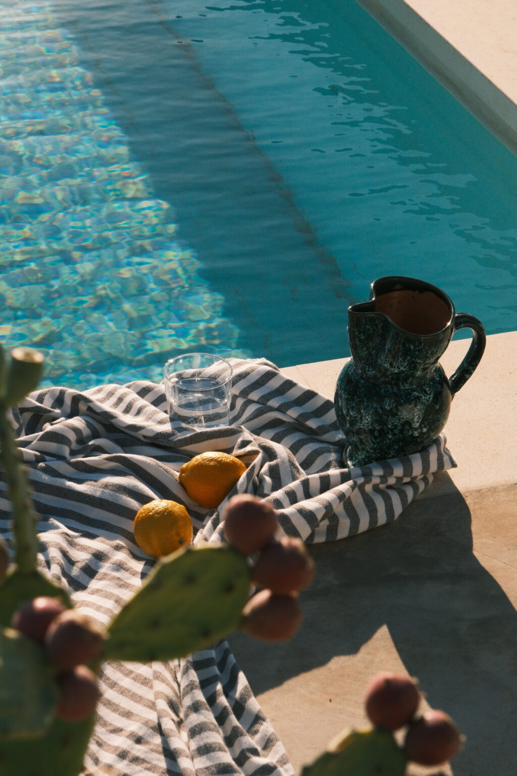 A serene poolside setting featuring a striped towel, a green ceramic pitcher, a glass of water, and two fresh lemons, with a cactus plant in the foreground. The crystal-clear water reflects sunlight, creating a tranquil atmosphere perfect for relaxation. | Only on SopranoVillas