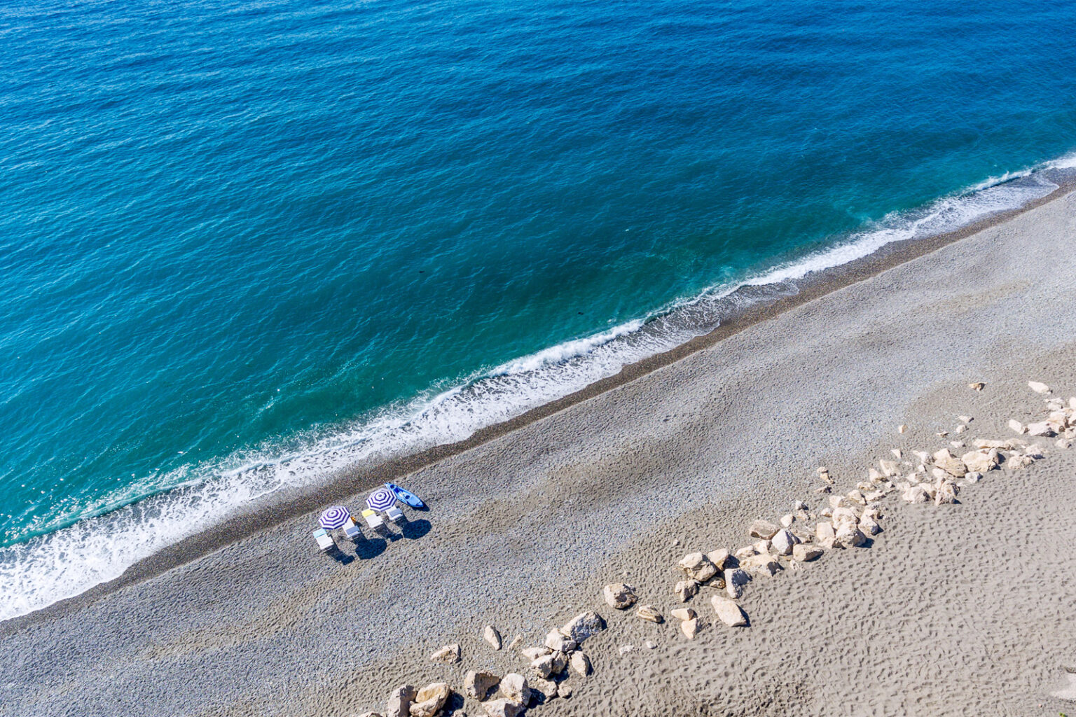 Aerial view of a serene beach with turquoise water, soft waves, and a pebbly shoreline, featuring beach umbrellas and lounge chairs for relaxation. | Only on SopranoVillas