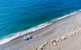 Aerial view of a serene beach with turquoise water, soft waves, and a pebbly shoreline, featuring beach umbrellas and lounge chairs for relaxation.