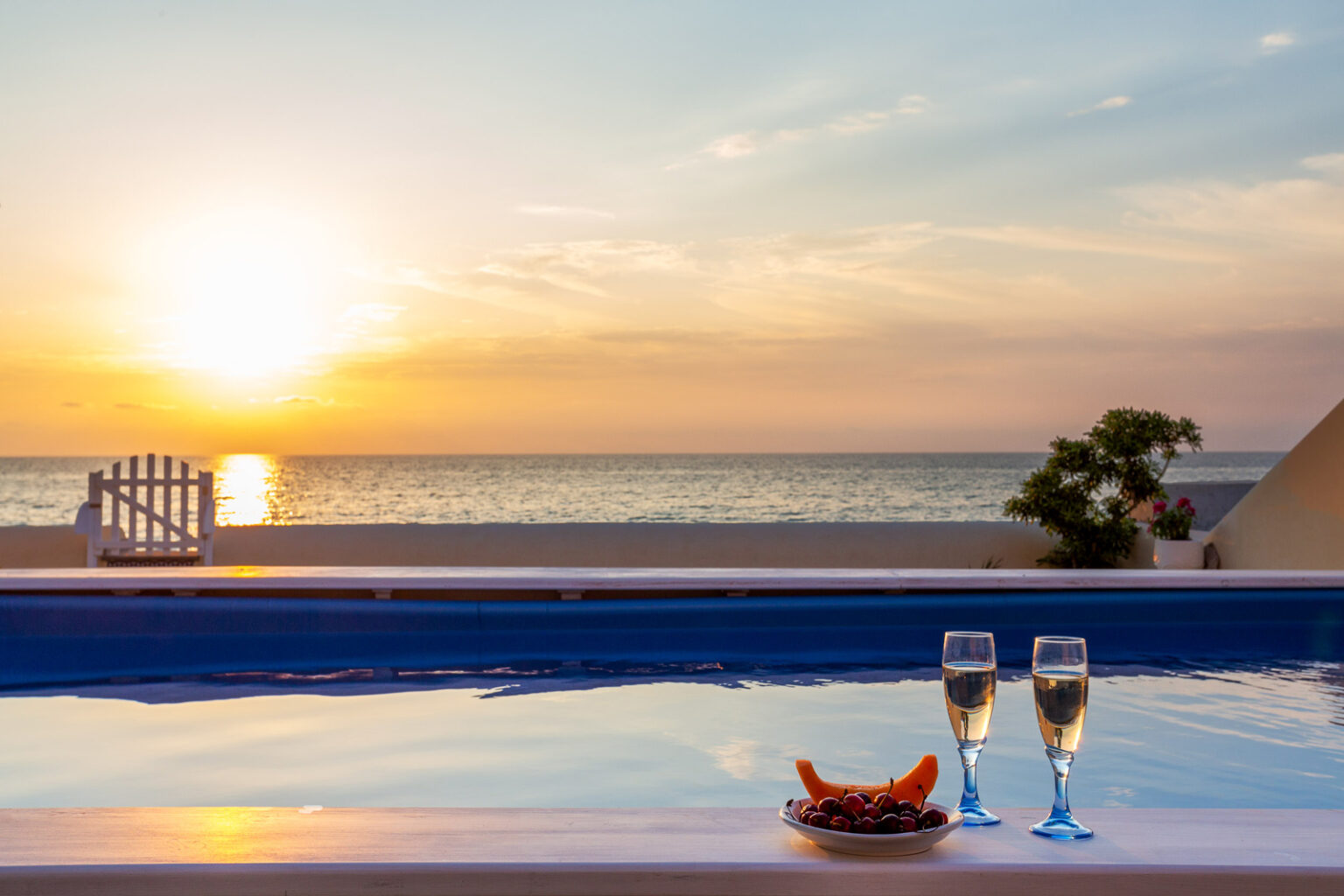 Sunset over a tranquil beach with a swimming pool in the foreground, featuring two glasses of champagne and a plate of fruit, creating a romantic and relaxing atmosphere. | Only on SopranoVillas
