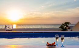 Sunset over a tranquil beach with a swimming pool in the foreground, featuring two glasses of champagne and a plate of fruit, creating a romantic and relaxing atmosphere.