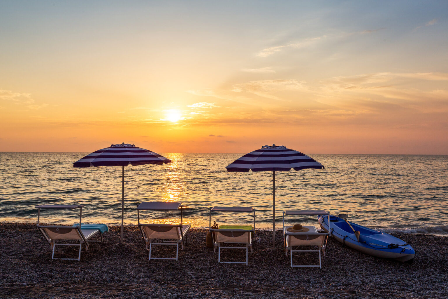 Beach scene at sunset featuring striped umbrellas and lounge chairs along the shoreline, with calm waves and a kayak nearby. Perfect for relaxation and summer vacations. | Only on SopranoVillas