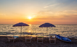 Beach scene at sunset featuring striped umbrellas and lounge chairs along the shoreline, with calm waves and a kayak nearby. Perfect for relaxation and summer vacations.