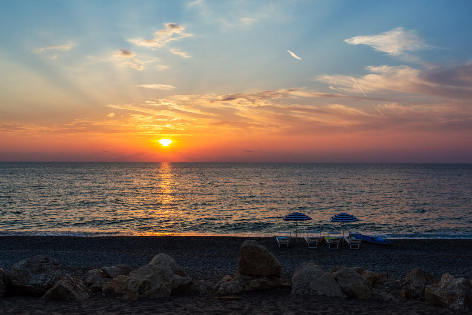 Sunset over the ocean with vibrant colors reflecting on the water, beach umbrellas in the foreground, and rocky shoreline. Ideal for travel and nature enthusiasts. | Only on SopranoVillas