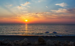 Sunset over the ocean with vibrant colors reflecting on the water, beach umbrellas in the foreground, and rocky shoreline. Ideal for travel and nature enthusiasts.